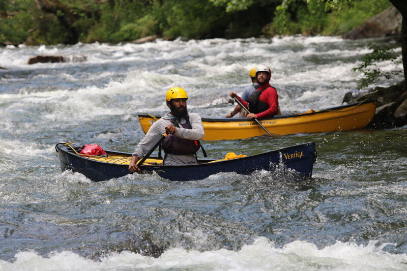 Two people are paddling canoes in a swiftly moving river. The person in the foreground canoe is wearing a yellow helmet and a gray long-sleeved shirt. The canoe is dark blue. The person in the background canoe is wearing a red shirt and a white helmet. The canoe is yellow. The river is turbulent with white water rapids.